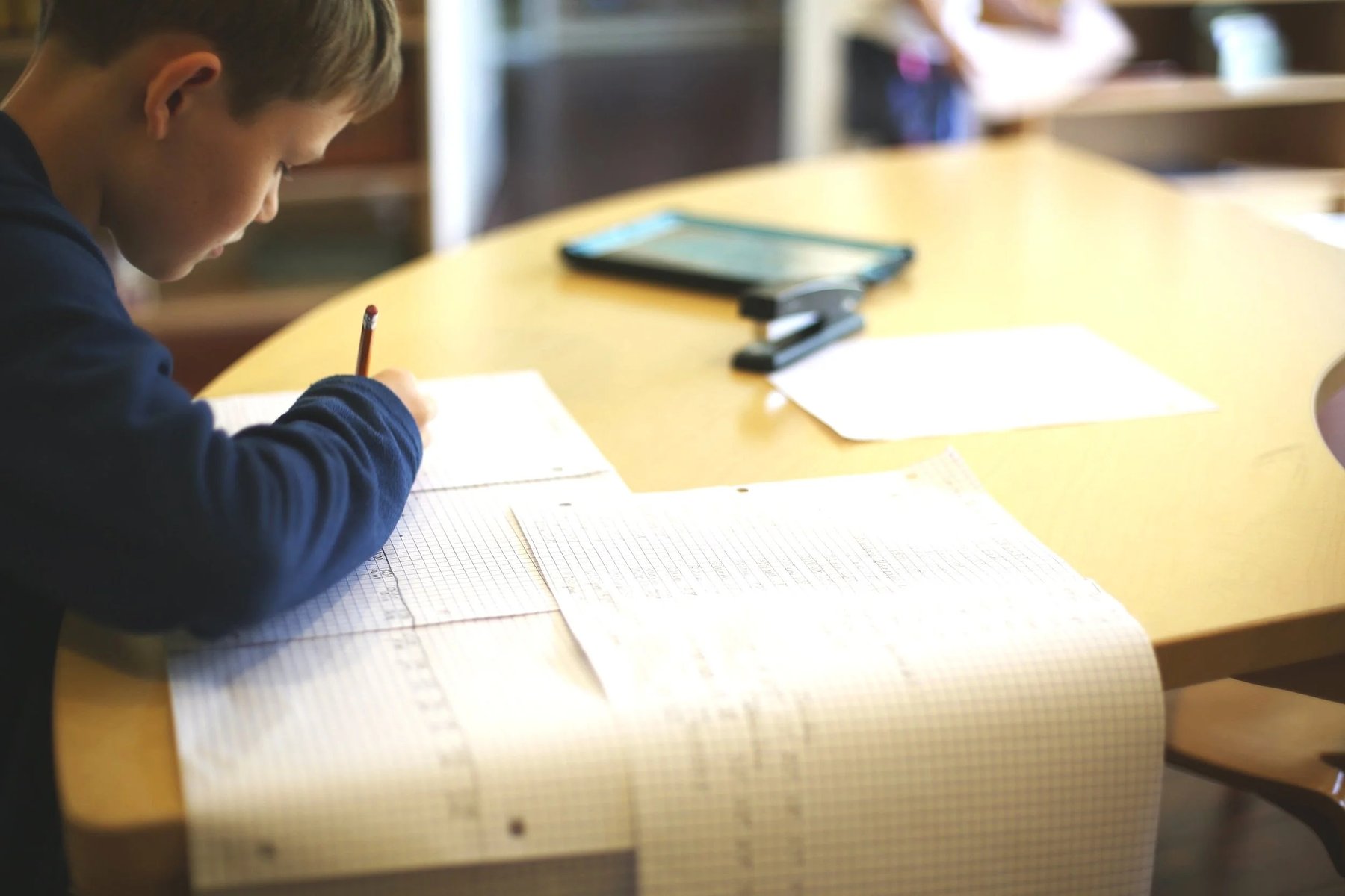 A young boy in a dark blue shirt writes with a pencil on a large sheet of grid paper at a wooden classroom table