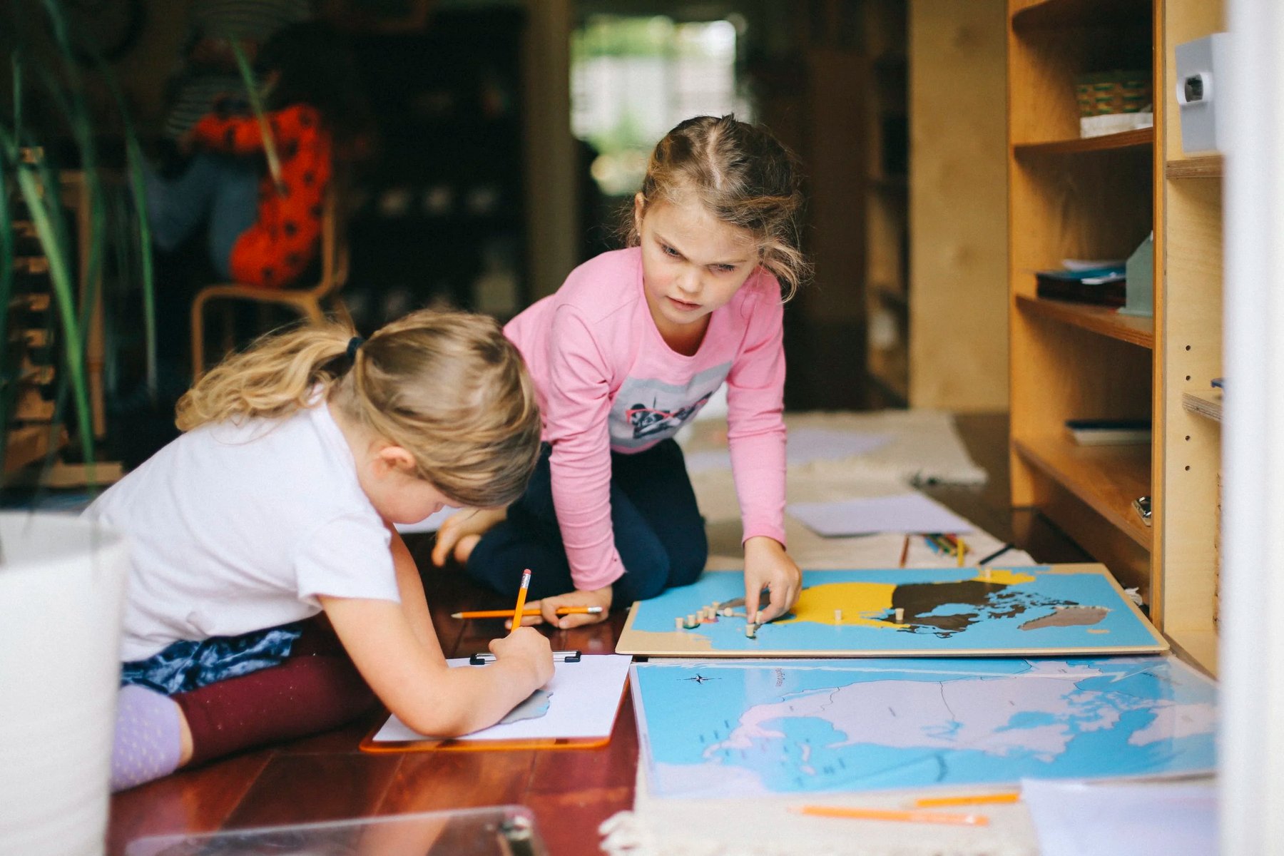 Two girls work on geography on the classroom floor: one assembles a wooden puzzle map of North America while the other writes notes beside a printed map