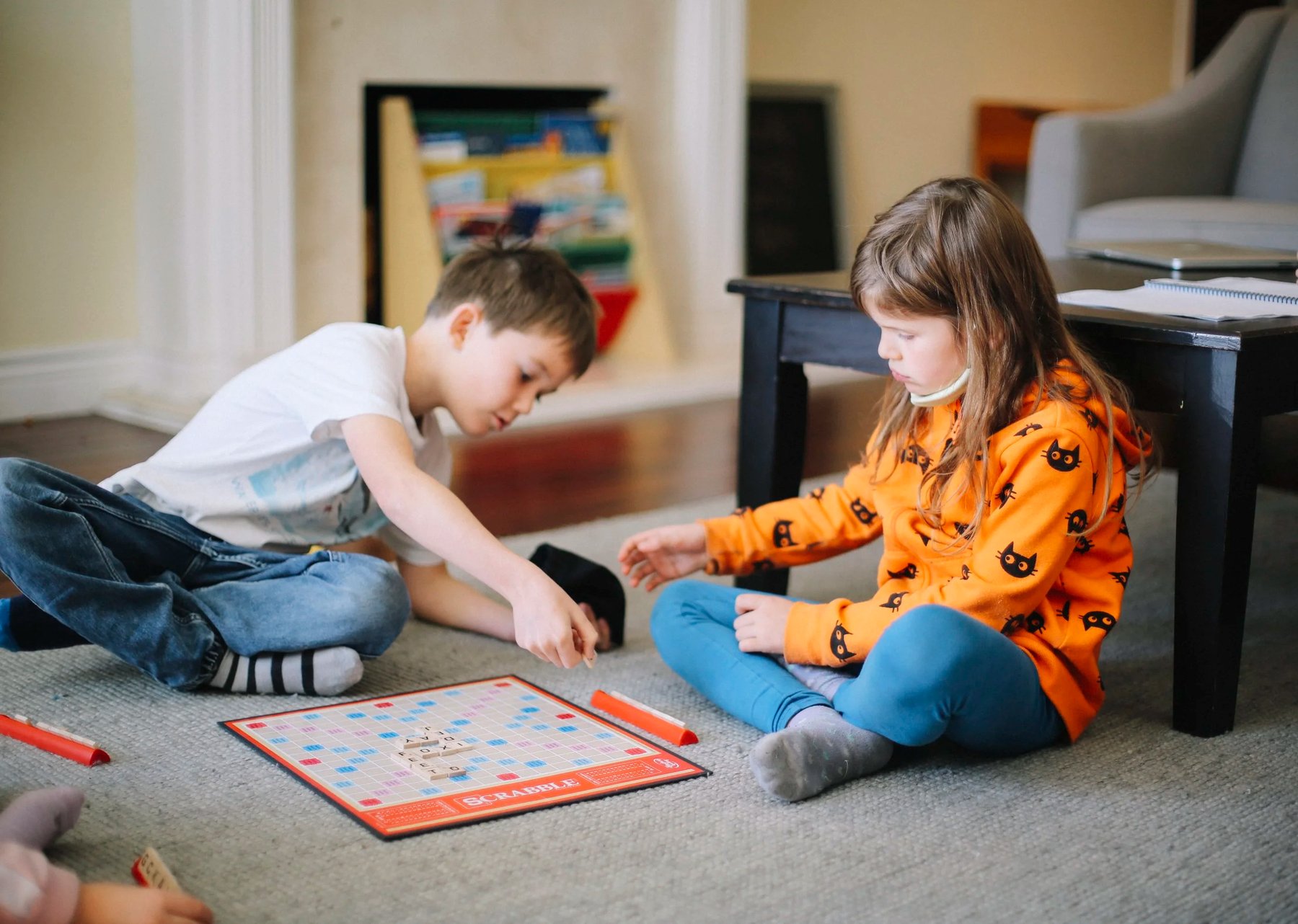 A boy and a girl sit on a carpet playing Scrabble with words formed on the board between their tile racks