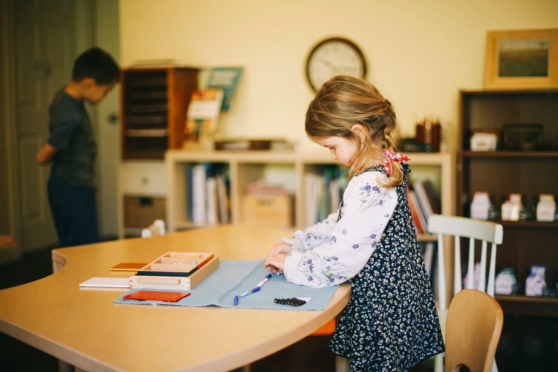 A girl with a blonde braid arranges small Montessori bead materials on a light blue mat at a classroom table