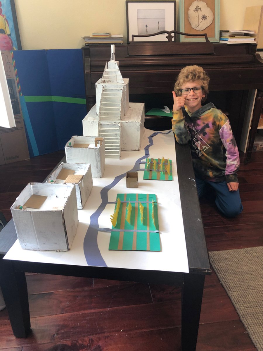 A boy in a tie-dye hoodie gives a thumbs-up beside his tabletop model of an ancient city with a stepped pyramid and a blue paper river