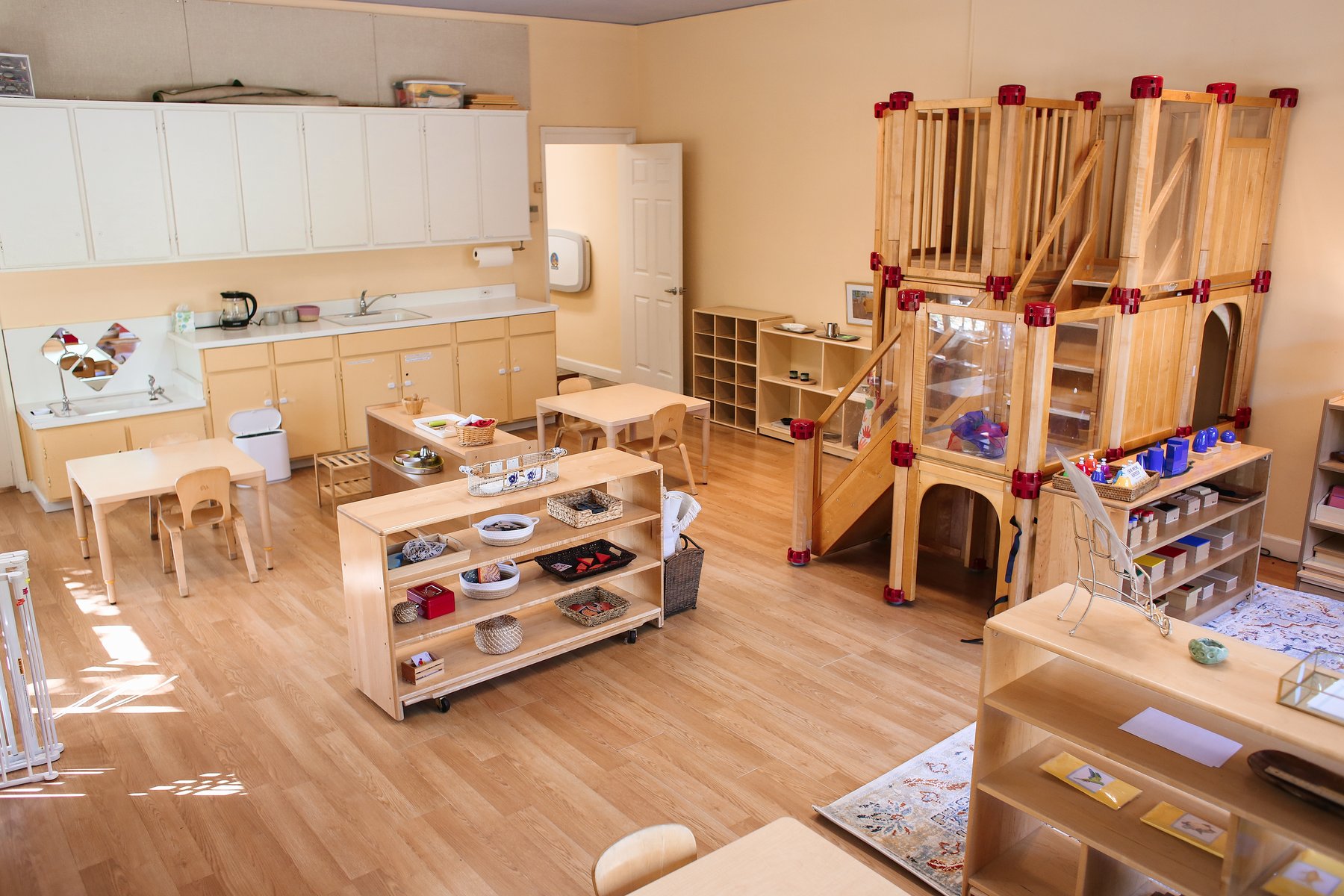 Wide early-childhood Montessori classroom with light wood floors, child-height kitchenette, low shelves of materials, and a multi-level wooden climbing structure