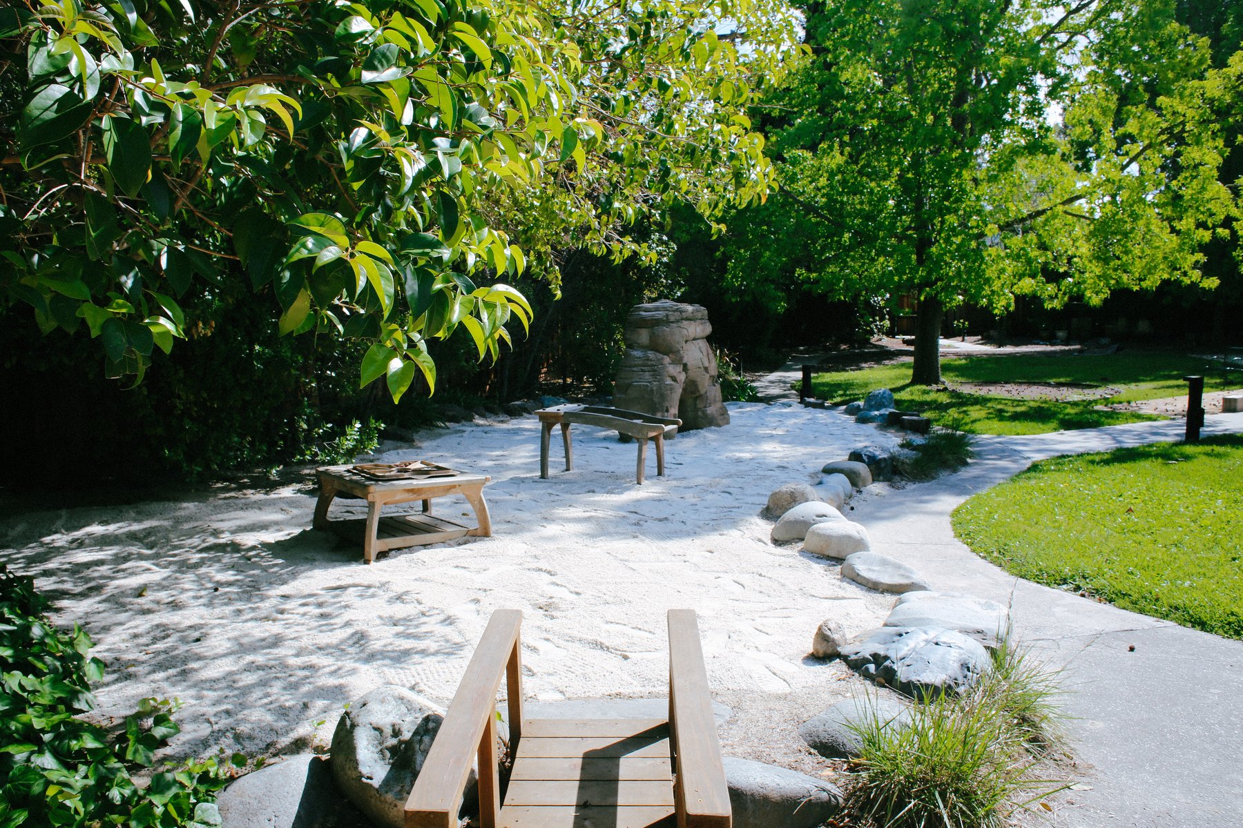 Sunny outdoor sand play area with a wooden footbridge, smooth-stone borders, low platform, and a climbing boulder beneath shade trees