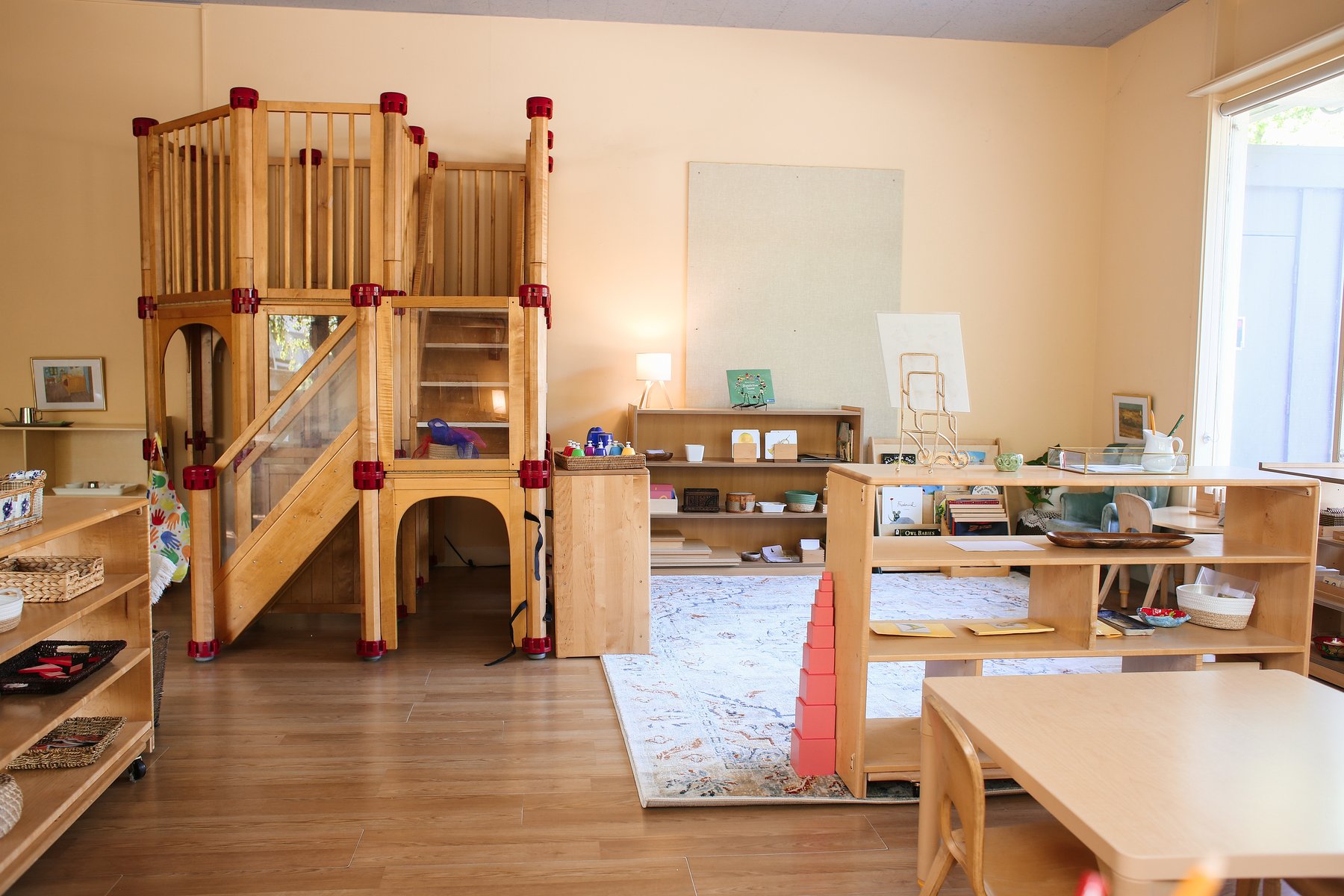 Bright preschool classroom with a two-level wooden indoor play structure across from low Montessori shelves stocked with materials including a Pink Tower