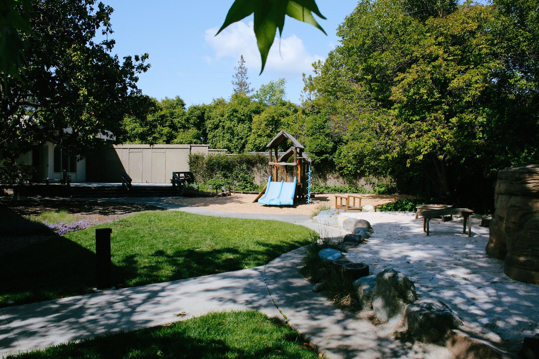 Sunny outdoor playground with a central wooden structure, blue double slide, sandpit, and curving paths beside the school building