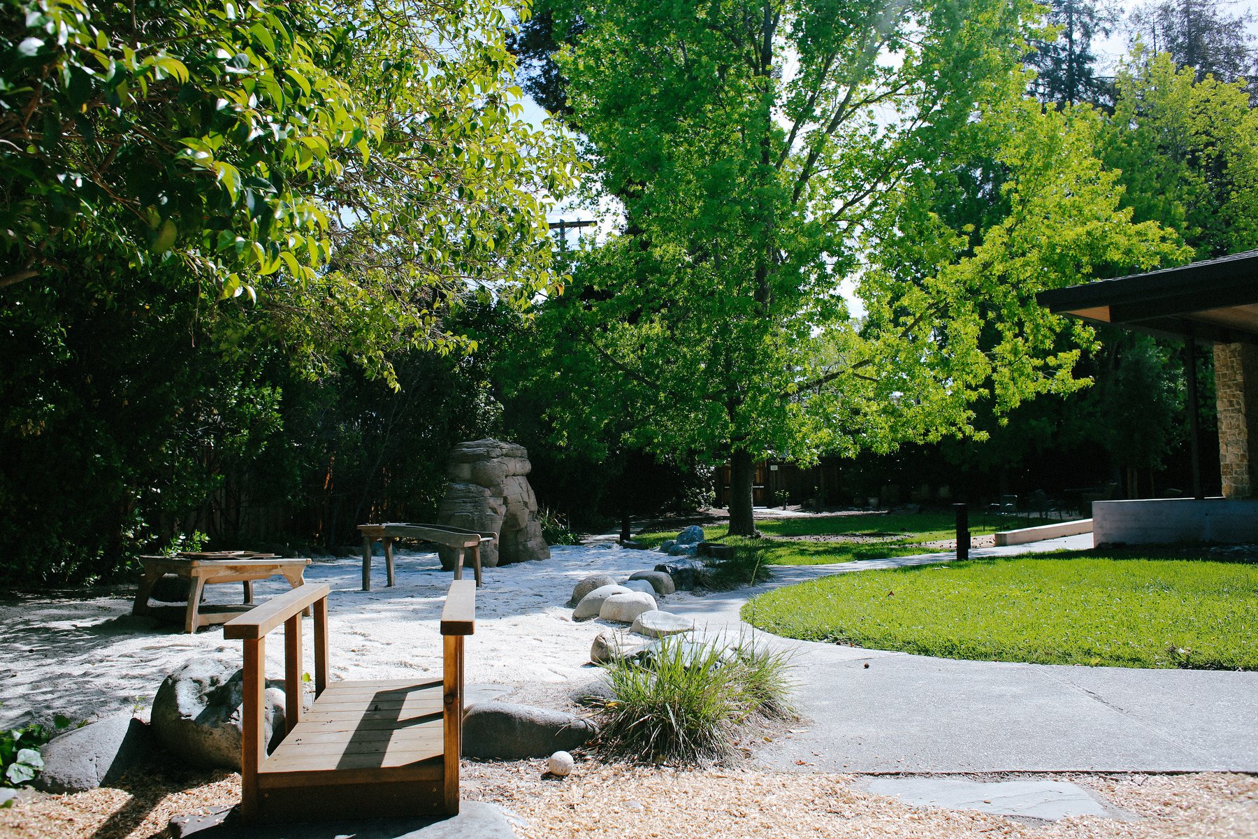 Naturalistic schoolyard with a small wooden bridge into a sandy play area edged by river rocks, low tables, and a faux-rock climber under leafy trees