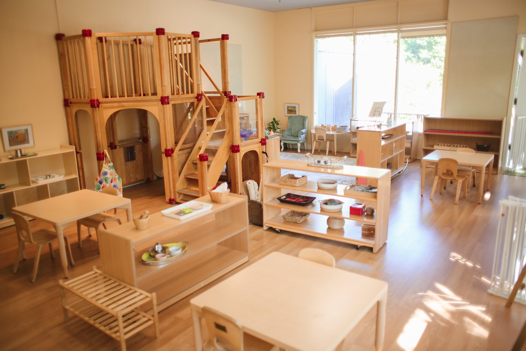 Spacious early-childhood classroom with a two-story wooden play loft, low shelves, child tables, and a teal armchair beneath tall sunlit windows