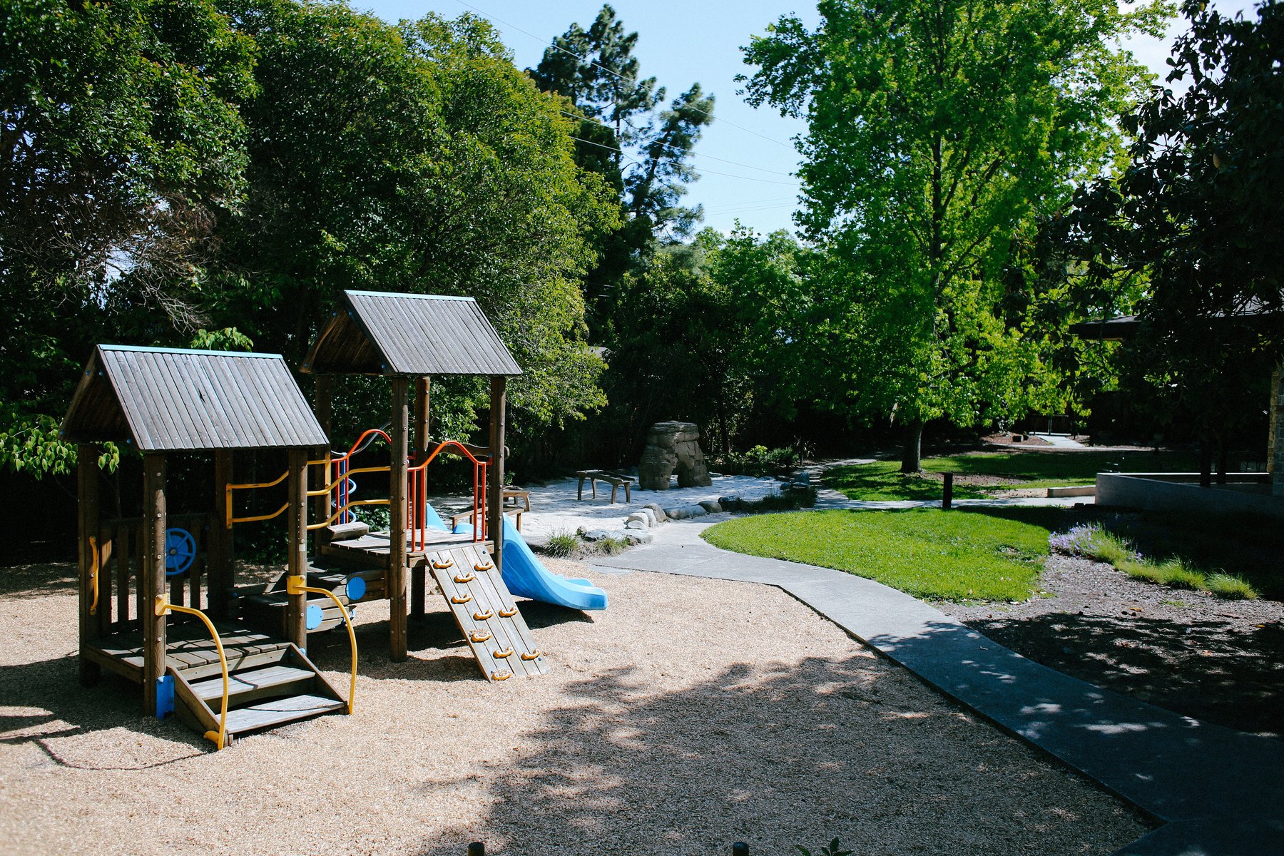 Colorful outdoor wooden play structure with twin towers, a blue slide, climbing ramp, and bright safety rails over wood-chip ground beside a sandy play area