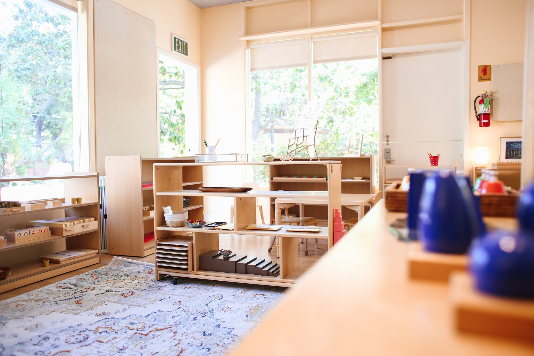 Sun-filled Montessori room with pale yellow walls, large windows onto trees, and low wooden shelves of blocks, beads, and trays around a patterned area rug