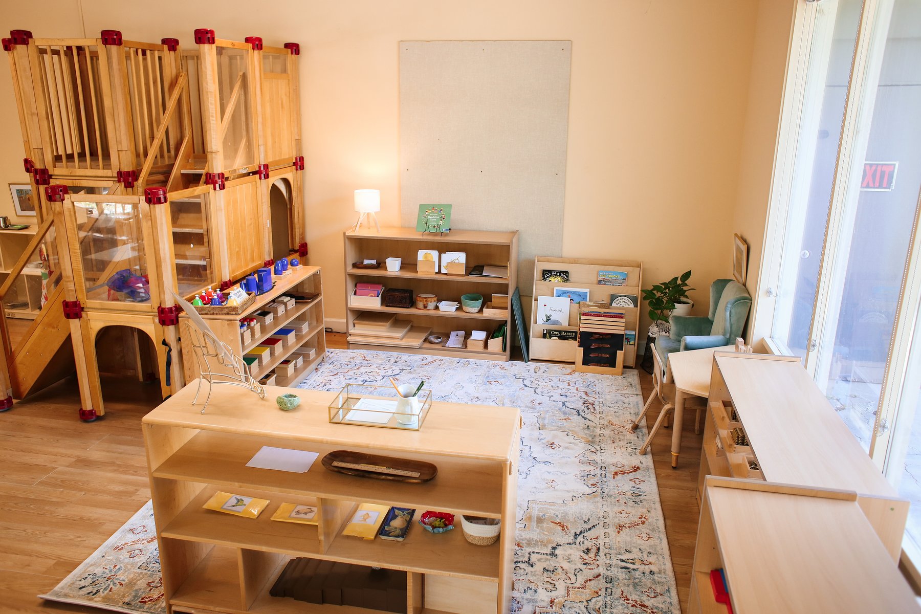 Preschool classroom with a large vintage-style rug, a two-tier shelf divider holding trays and small materials, a climbing loft, book display, and teal reading chair