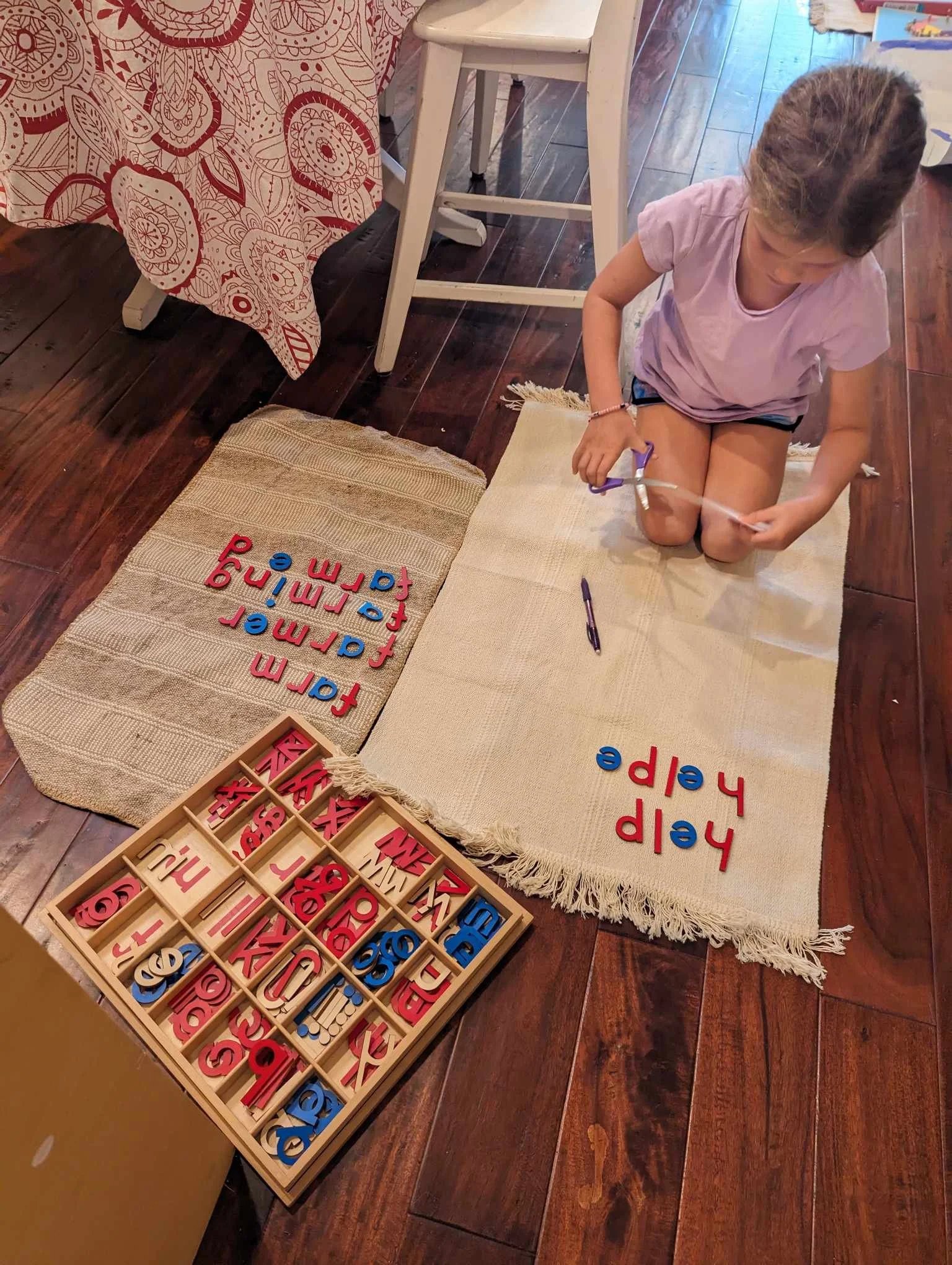 A girl kneels on hardwood placing movable alphabet words like "farm," "farmer," and "help" on woven rugs beside a segmented letter tray