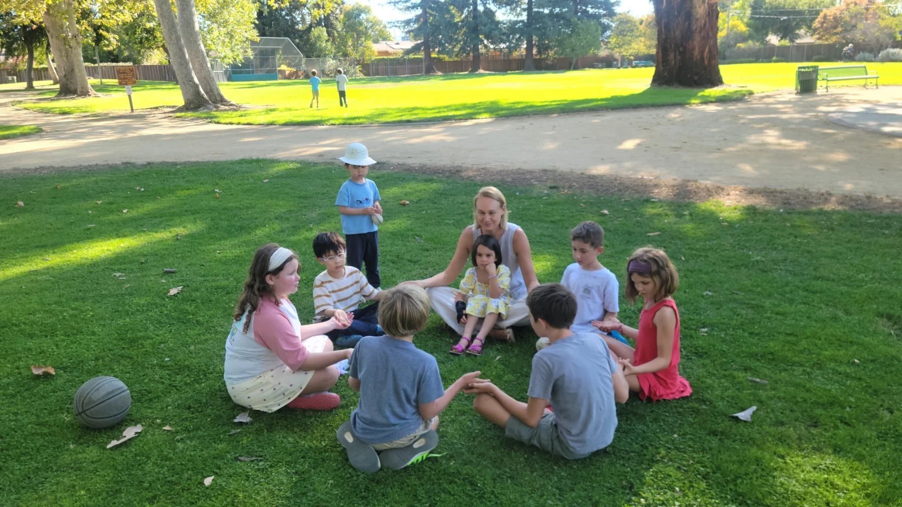 A teacher and learners of mixed ages sit in a grassy outdoor circle under park trees during an outdoor lesson